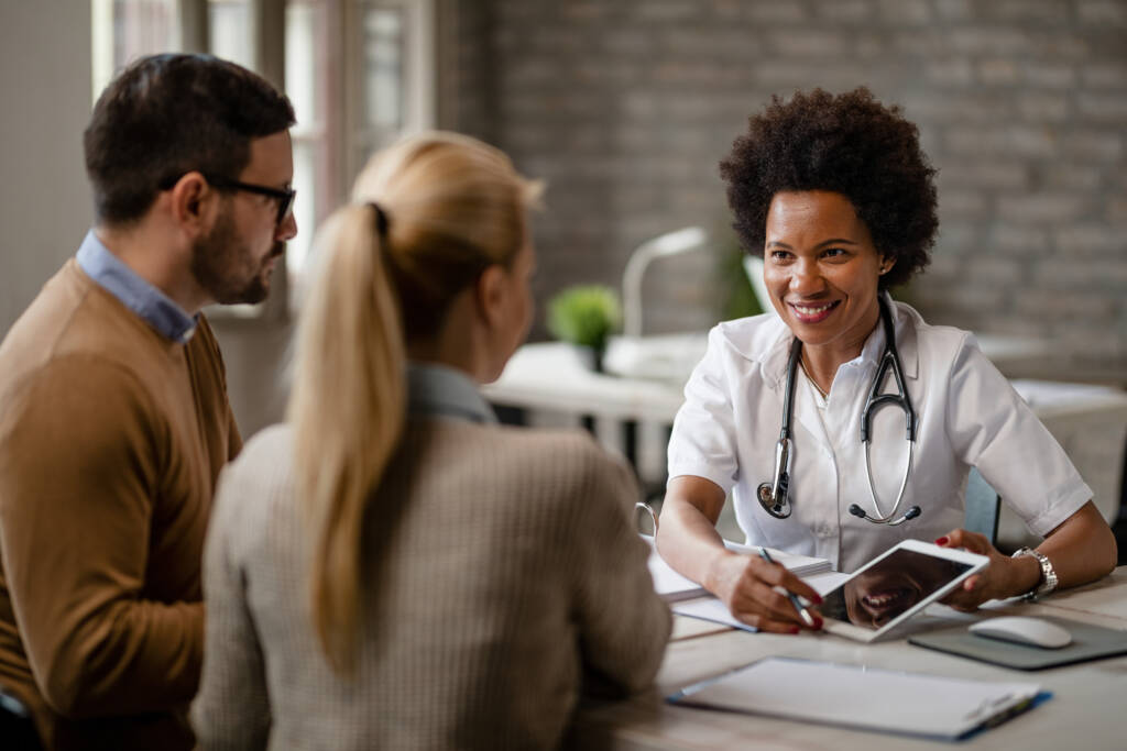 Happy black female doctor talking to a couple and showing them medical test results on a touchpad during consultations at clinic.